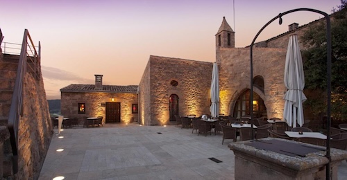 Foto del exterior del Parador de Cardona donde se puede ver las mesas exteriores del restaurante y los edificios de piedra al fondo.