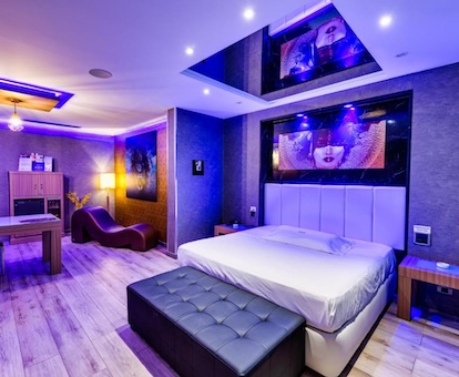 Habitación con decoración para adultos, luces fluorescentes, un sillón tantra al fondo y un espejo en el techo sobre la cama.